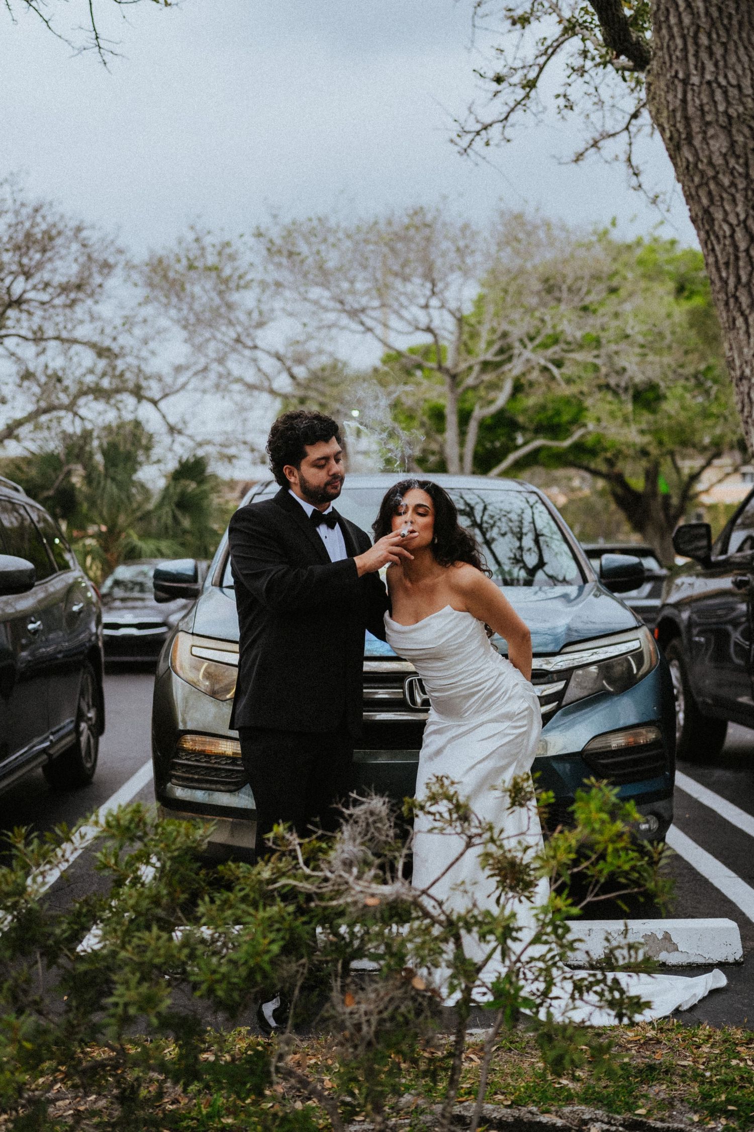 A couple in formal wedding attire shares a romantic moment between parked cars in an outdoor setting.