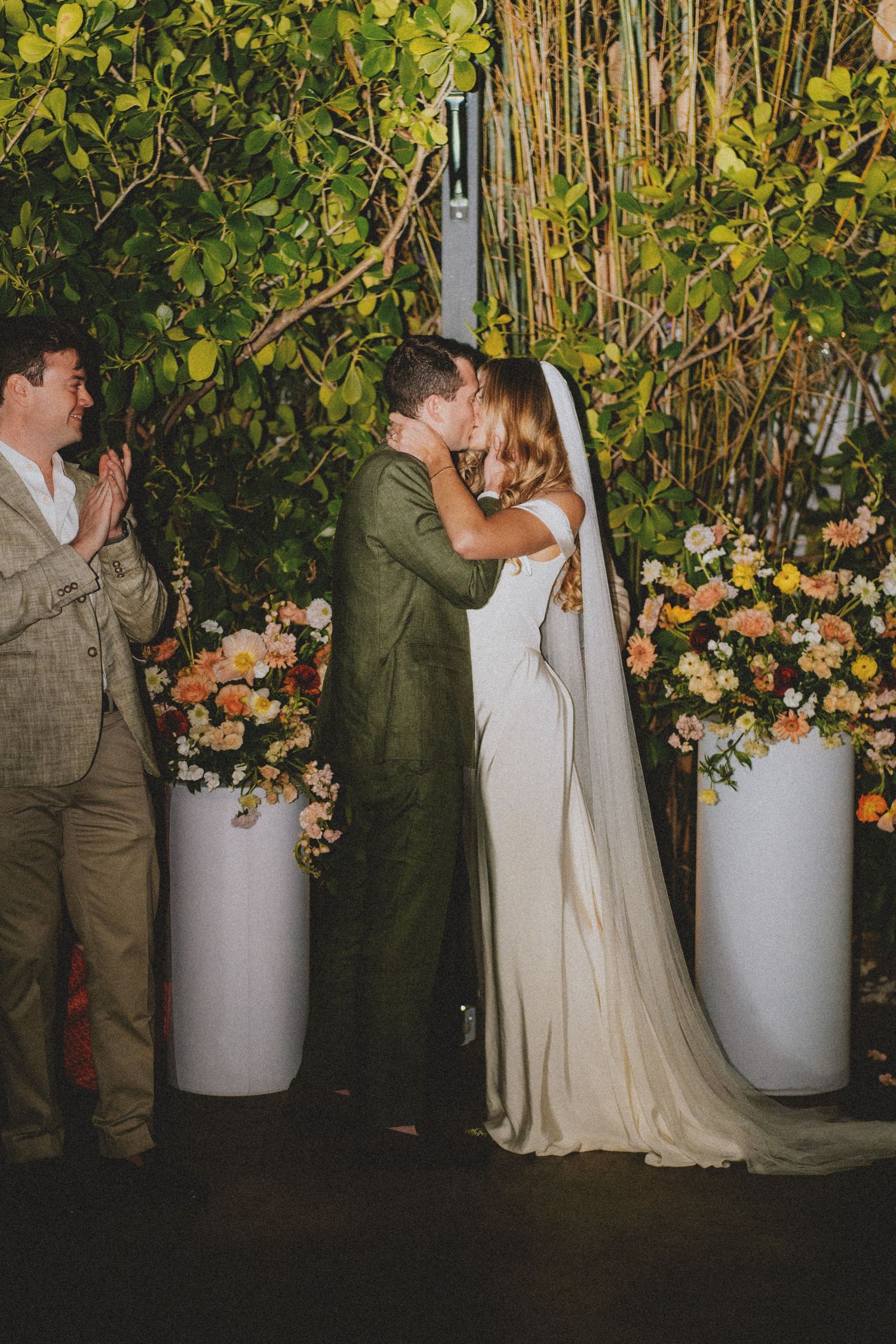 Bride and groom share first kiss at outdoor wedding ceremony with colorful floral arrangements and lush greenery backdrop.