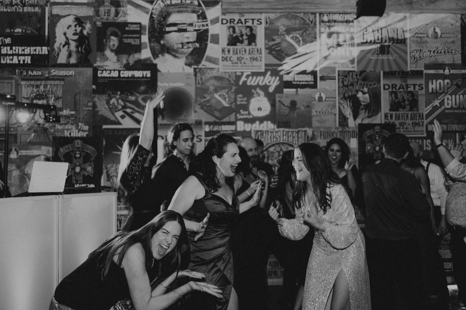 Black and white photo of women laughing and dancing at a lively party with vintage posters on the wall.