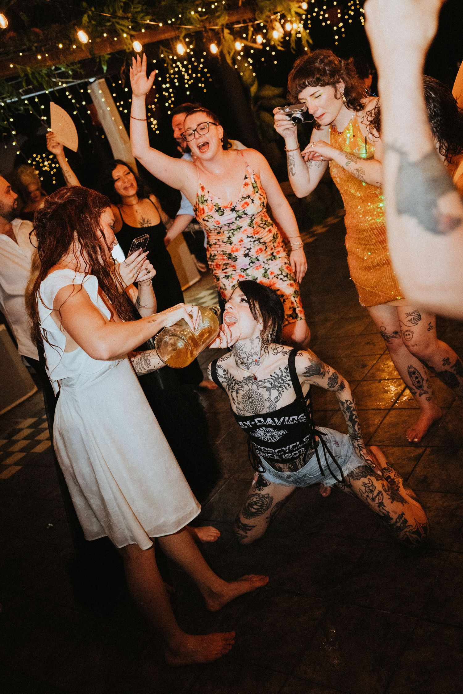 Guests dancing energetically at a lively outdoor wedding reception with string lights and tattoos visible.