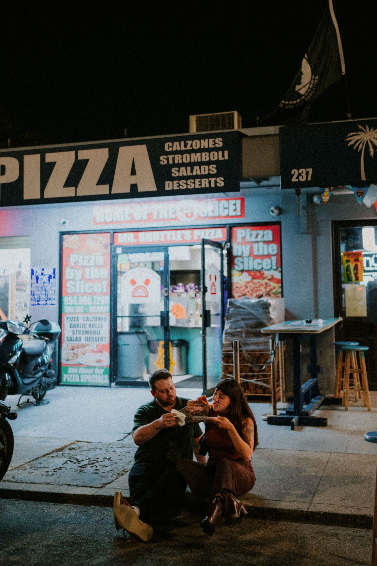 People sitting on sidewalk outside a pizza restaurant at night with illuminated storefront signage in the background.