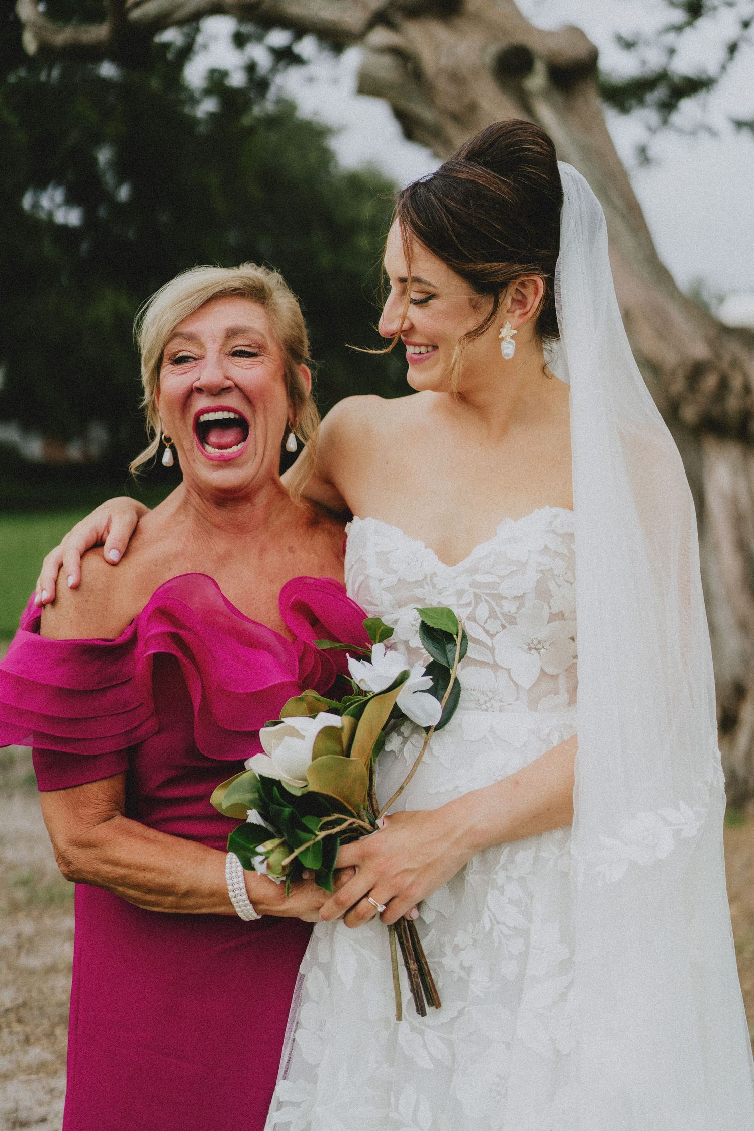Bride in white lace gown holding bouquet laughs with older woman in fuchsia ruffled dress at outdoor wedding.