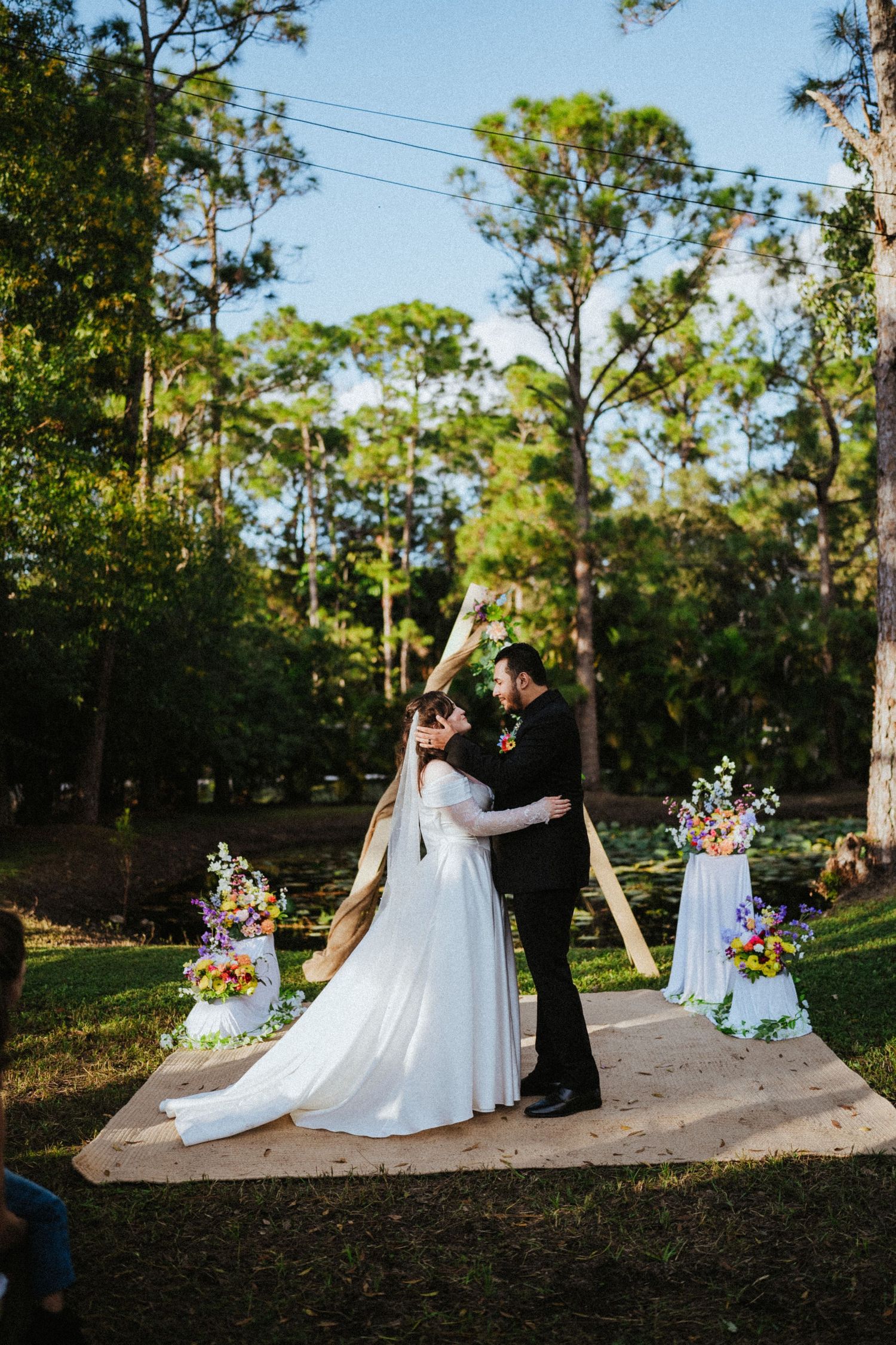 Bride and groom share first kiss at outdoor wedding ceremony with floral arch and tall pine trees in background.