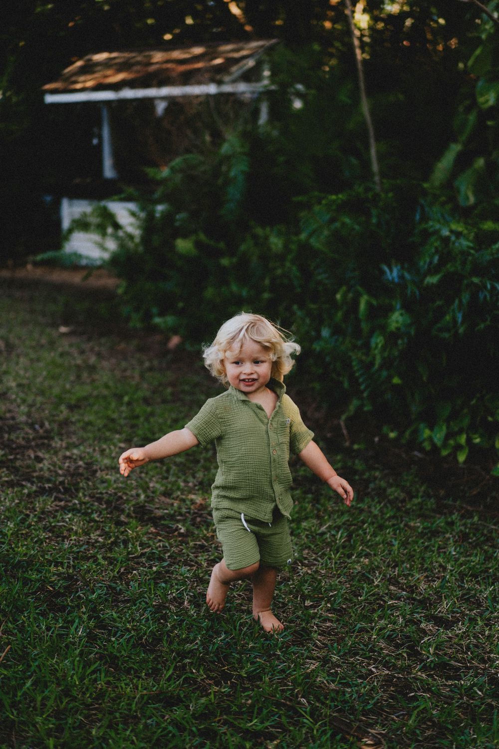 A child in a green outfit runs and plays joyfully on grass in a backyard setting.
