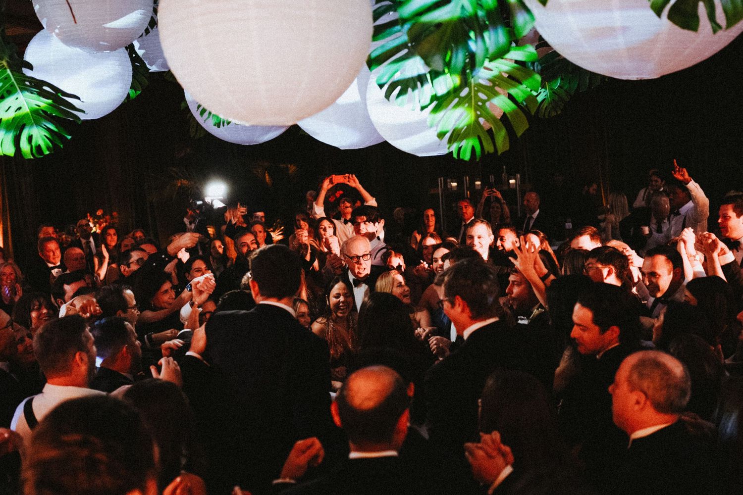 Crowded wedding reception dance floor with guests celebrating under white paper lantern lights at night.