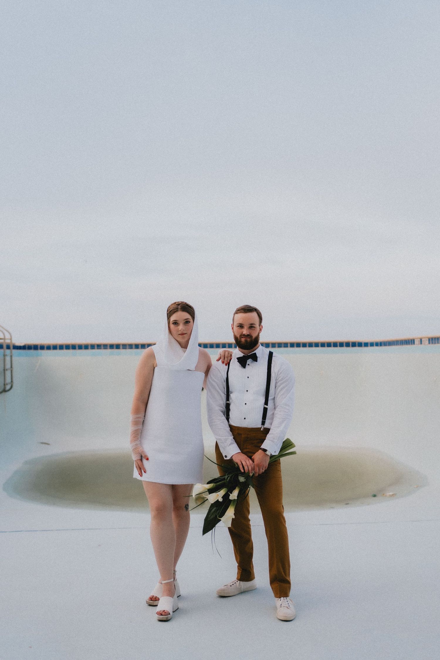 Bride in white hooded mini dress and groom in mustard pants pose by empty pool holding tropical bouquet.