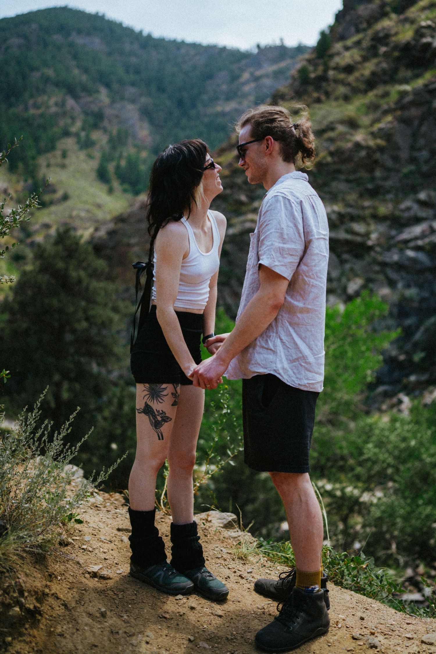 A couple holds hands on a rocky mountain trail, surrounded by lush green hills, dressed in casual hiking attire.