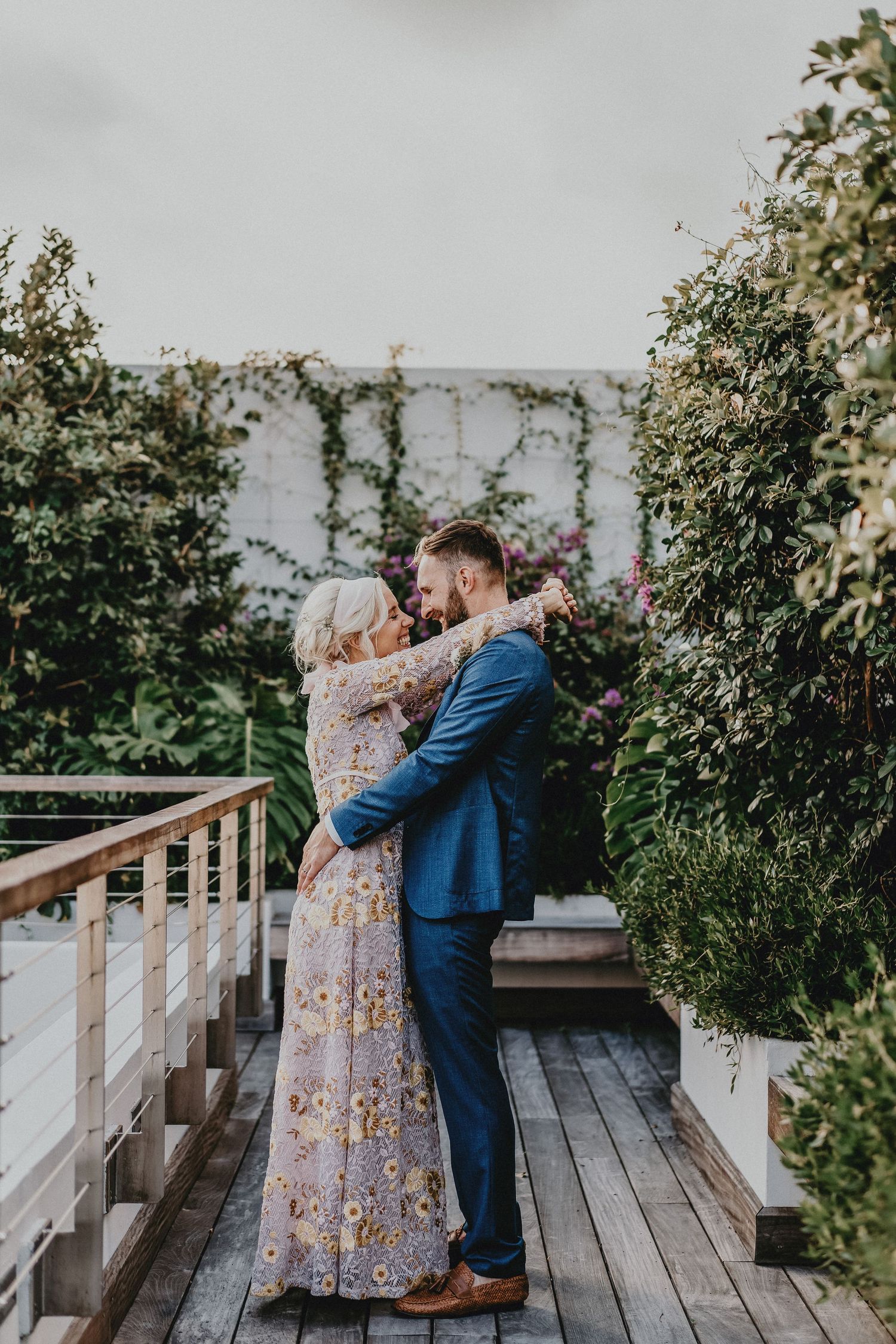 Couple embracing on a wooden deck surrounded by lush greenery, woman in floral gown and man in blue suit.