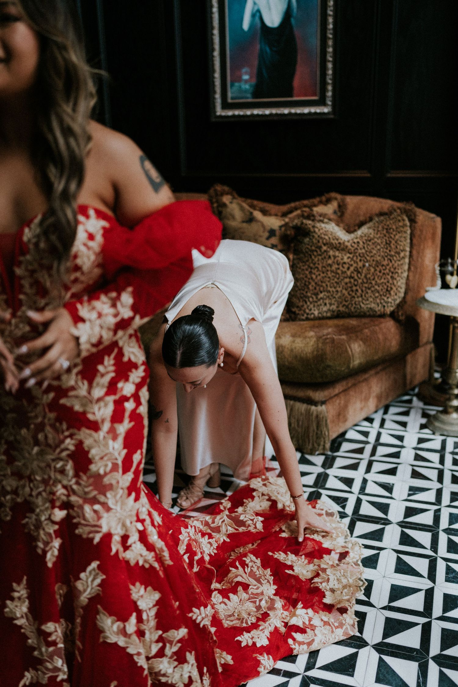 Bridesmaid in white slip dress adjusting red floral wedding gown train on black and white geometric floor.
