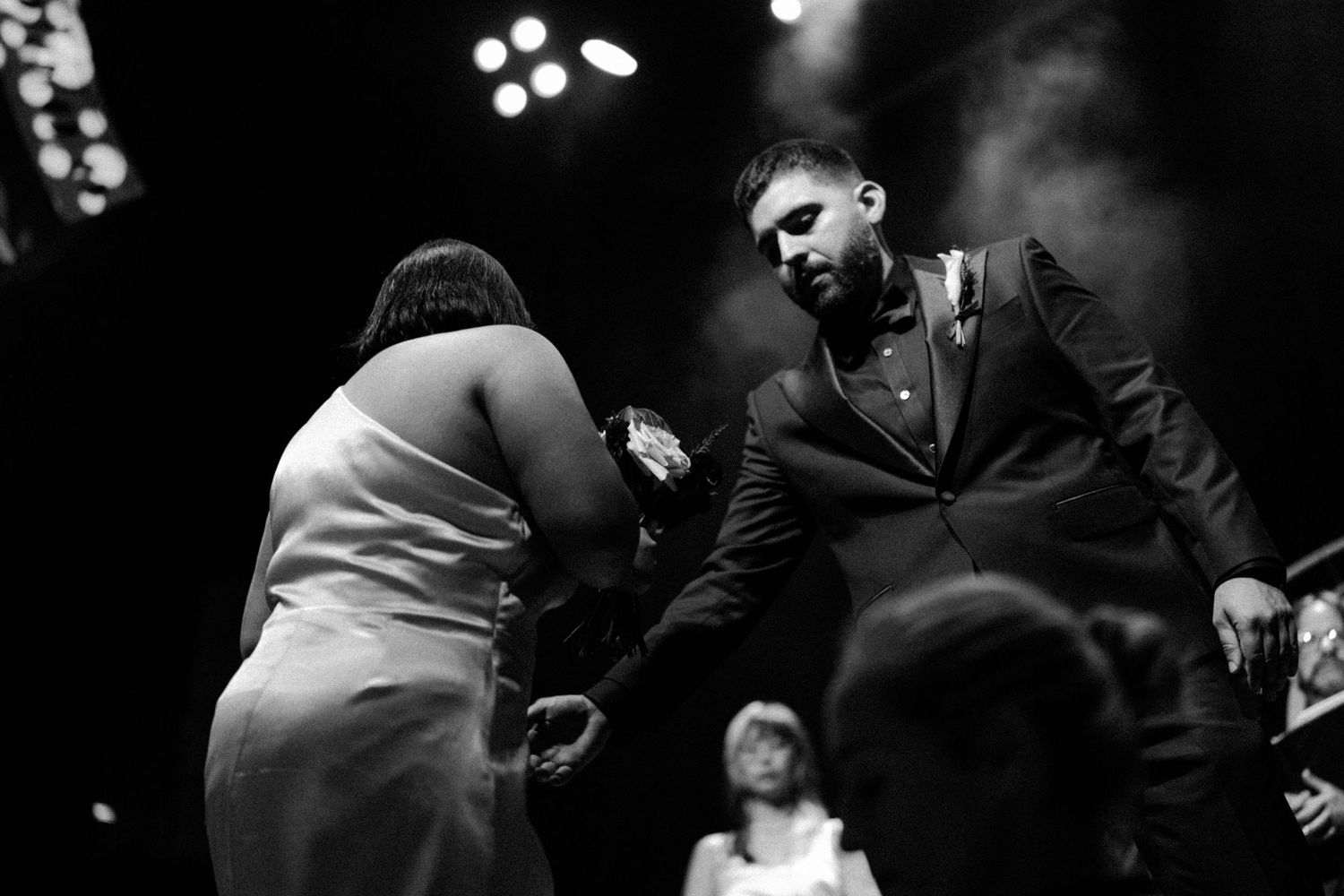Black and white photo of a groom in a suit bowing to hold hands with a bride in a white dress at a wedding reception.