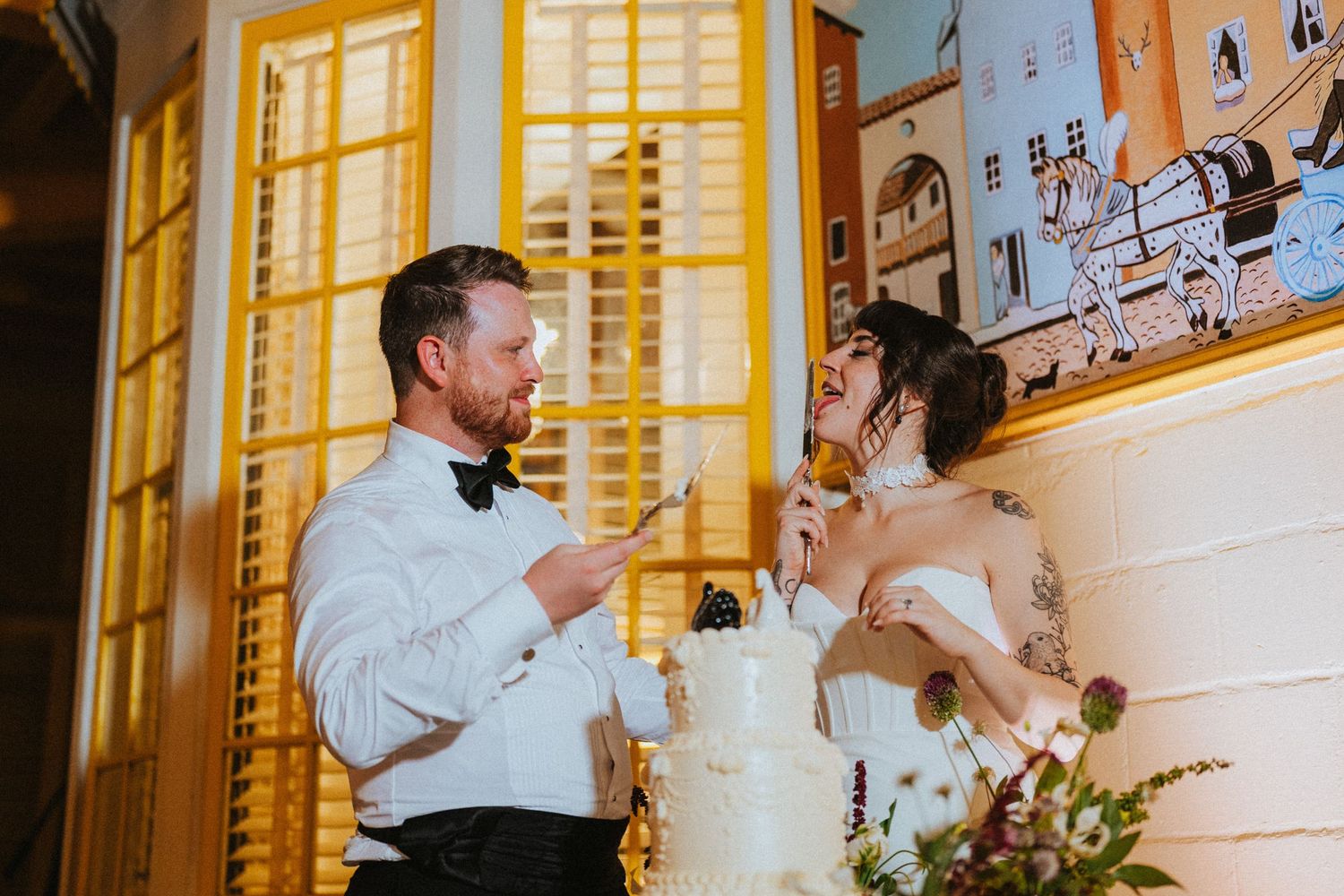 Bride and groom laughing while cutting their white wedding cake at a elegant indoor reception venue.