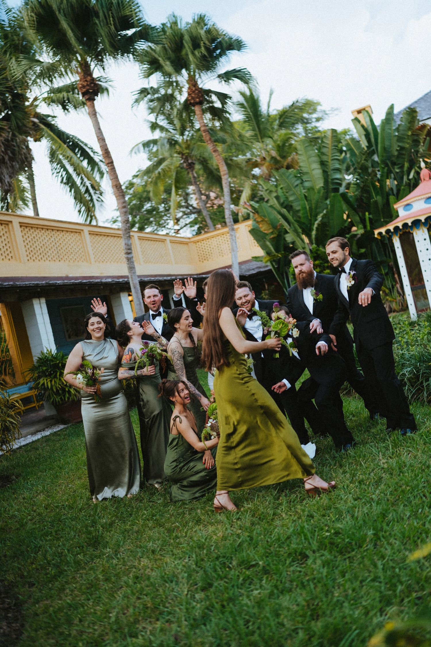 Bridal party in olive and gold dresses with groomsmen in tuxedos celebrating outdoors near tropical palm trees.
