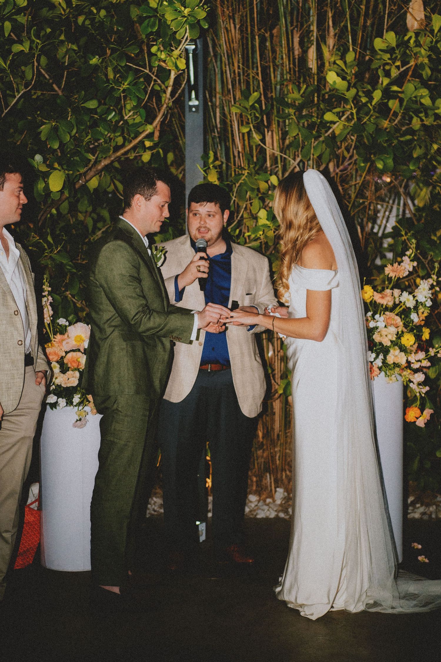 Couple exchanging wedding vows at outdoor ceremony with floral arrangements and lush greenery backdrop.