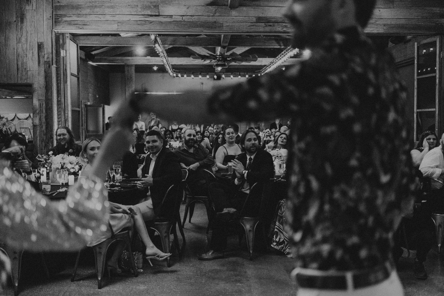 Black and white photo of wedding guests laughing and enjoying entertainment at a rustic barn reception.