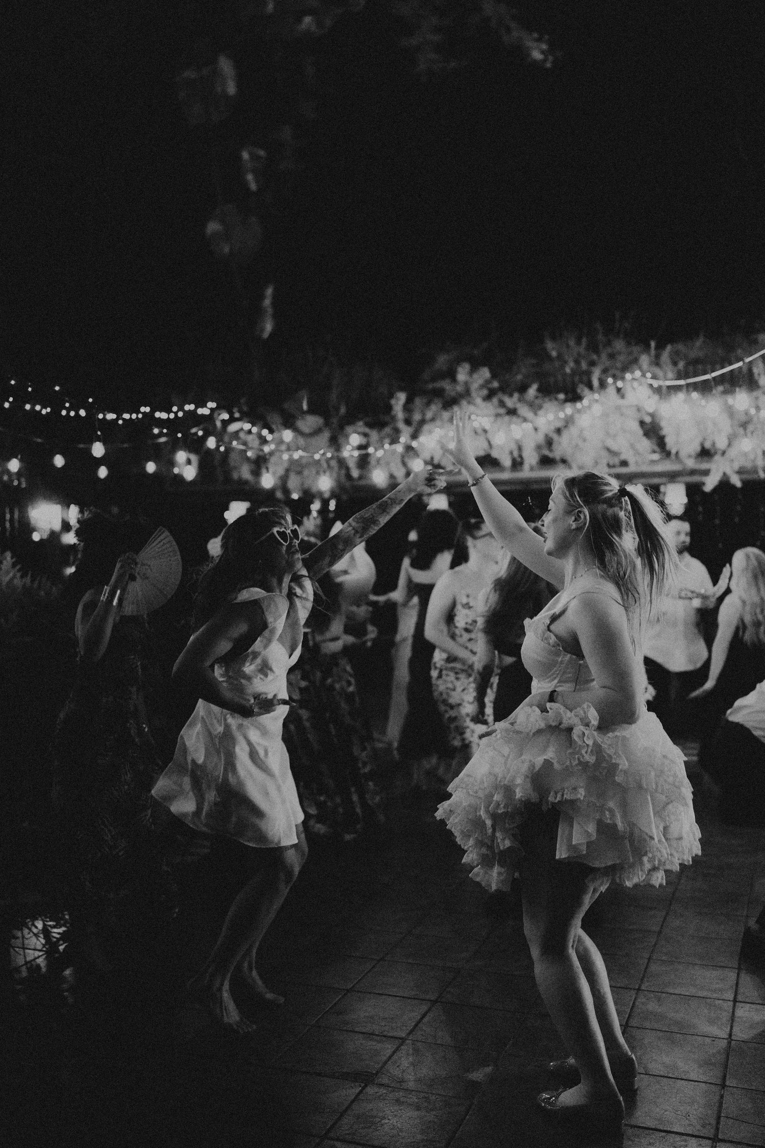 Black and white photo of women in white tutus dancing joyfully at an outdoor wedding reception at night.
