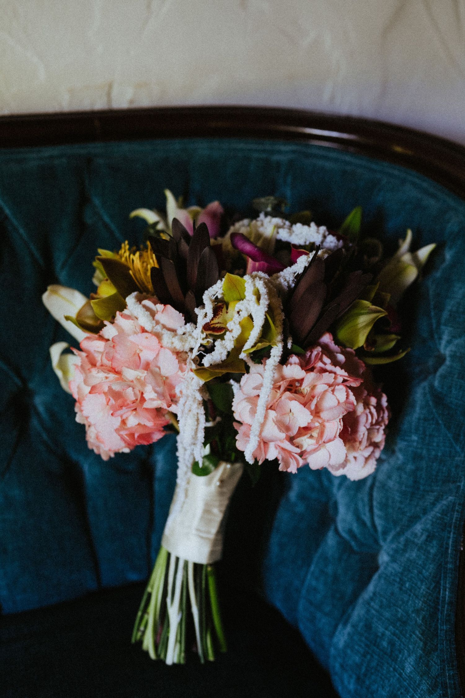 Pink hydrangea wedding bouquet with mixed blooms resting on teal velvet chair.