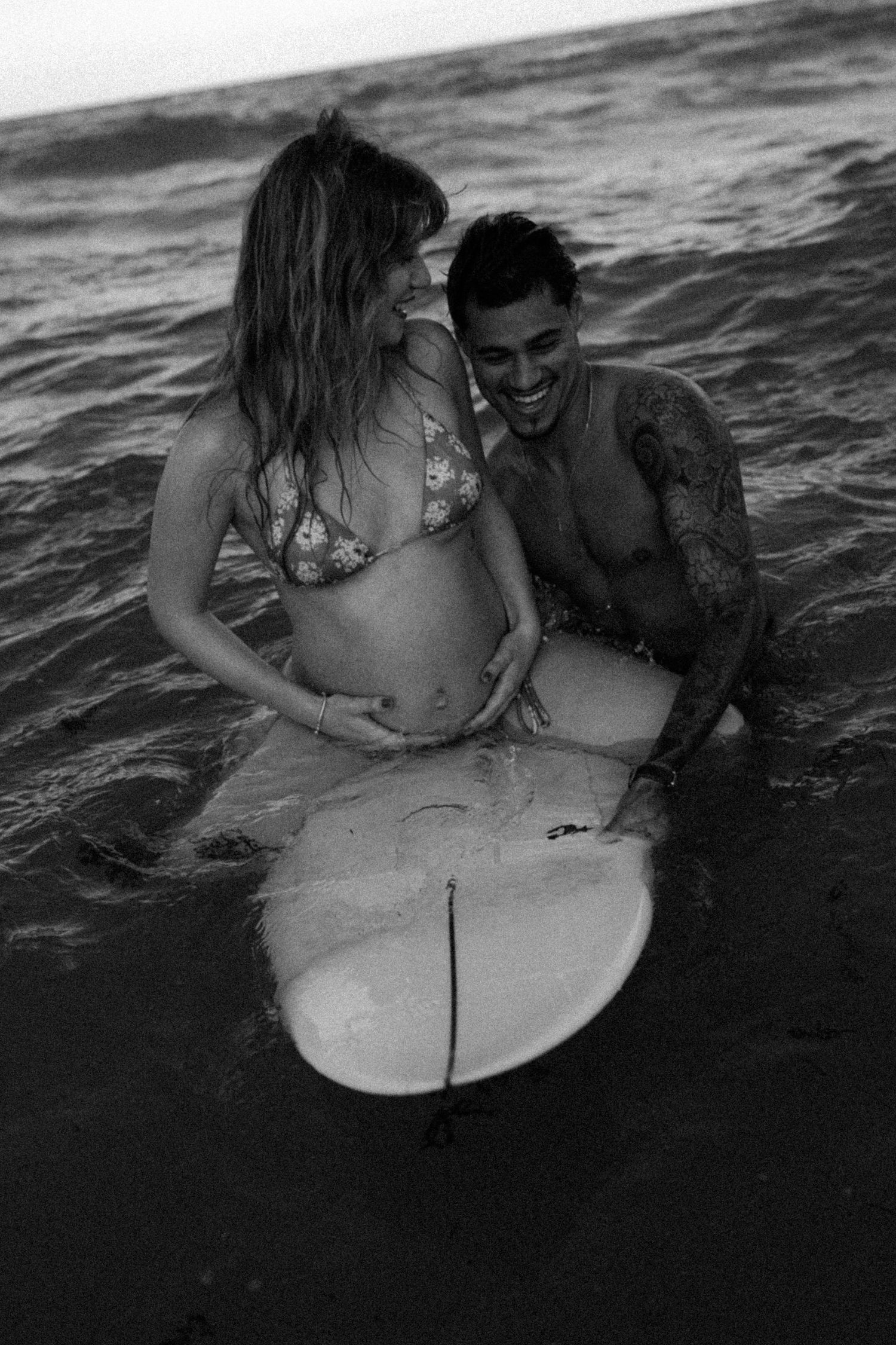 Pregnant couple smiling together in ocean waves with a surfboard in black and white beach maternity photo.