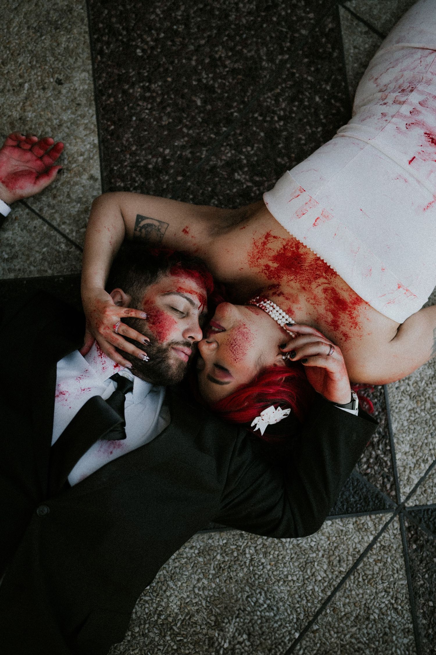 Couple in wedding attire covered in fake blood lying on ground for a dramatic zombie-themed Halloween wedding shoot.