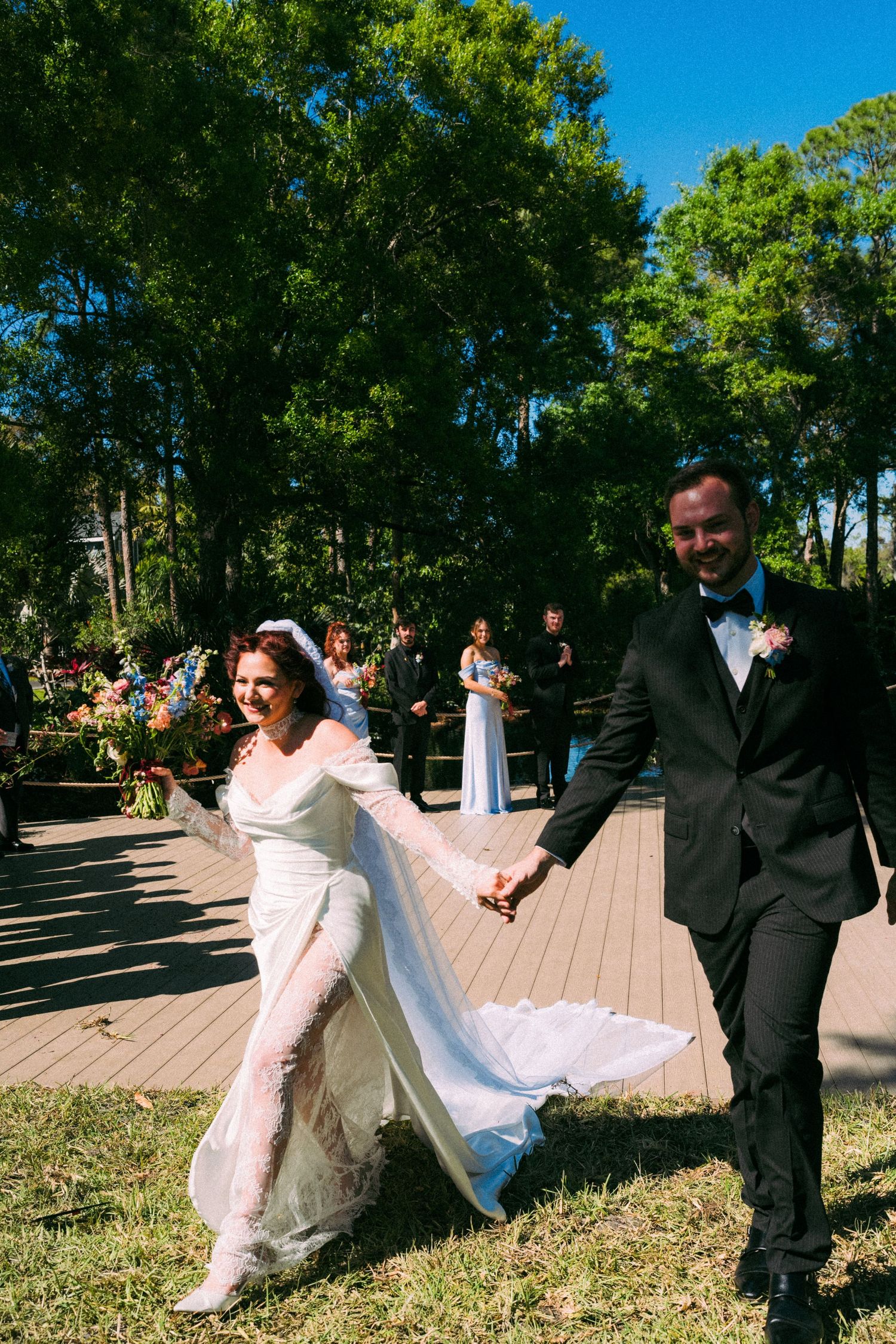 Bride in white gown and groom in black tuxedo walk hand-in-hand after outdoor wedding ceremony surrounded by lush green trees.