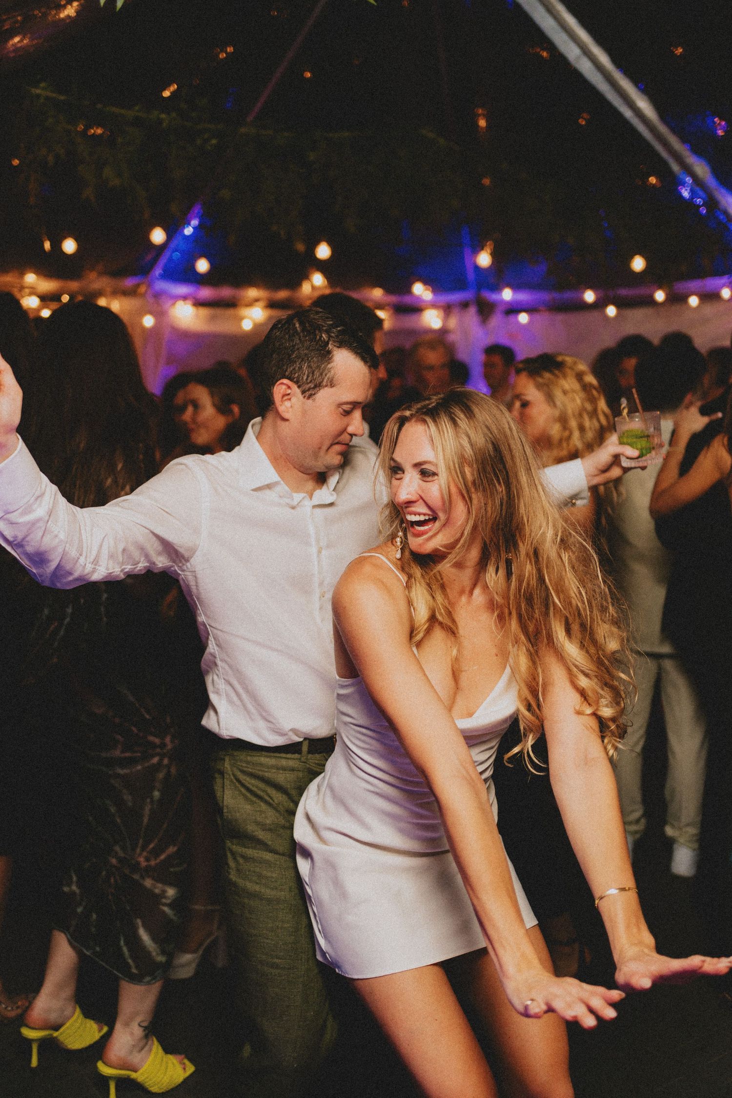 Couple dancing joyfully at a wedding reception under string lights in a tent.