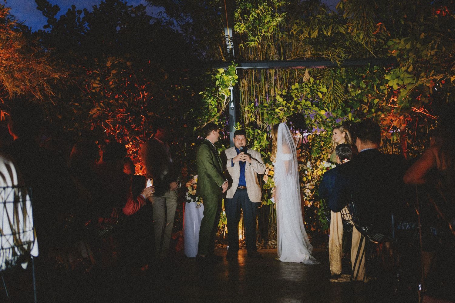 Outdoor nighttime wedding ceremony with bride and groom at floral arch, guests watching, surrounded by lush greenery and warm lighting.