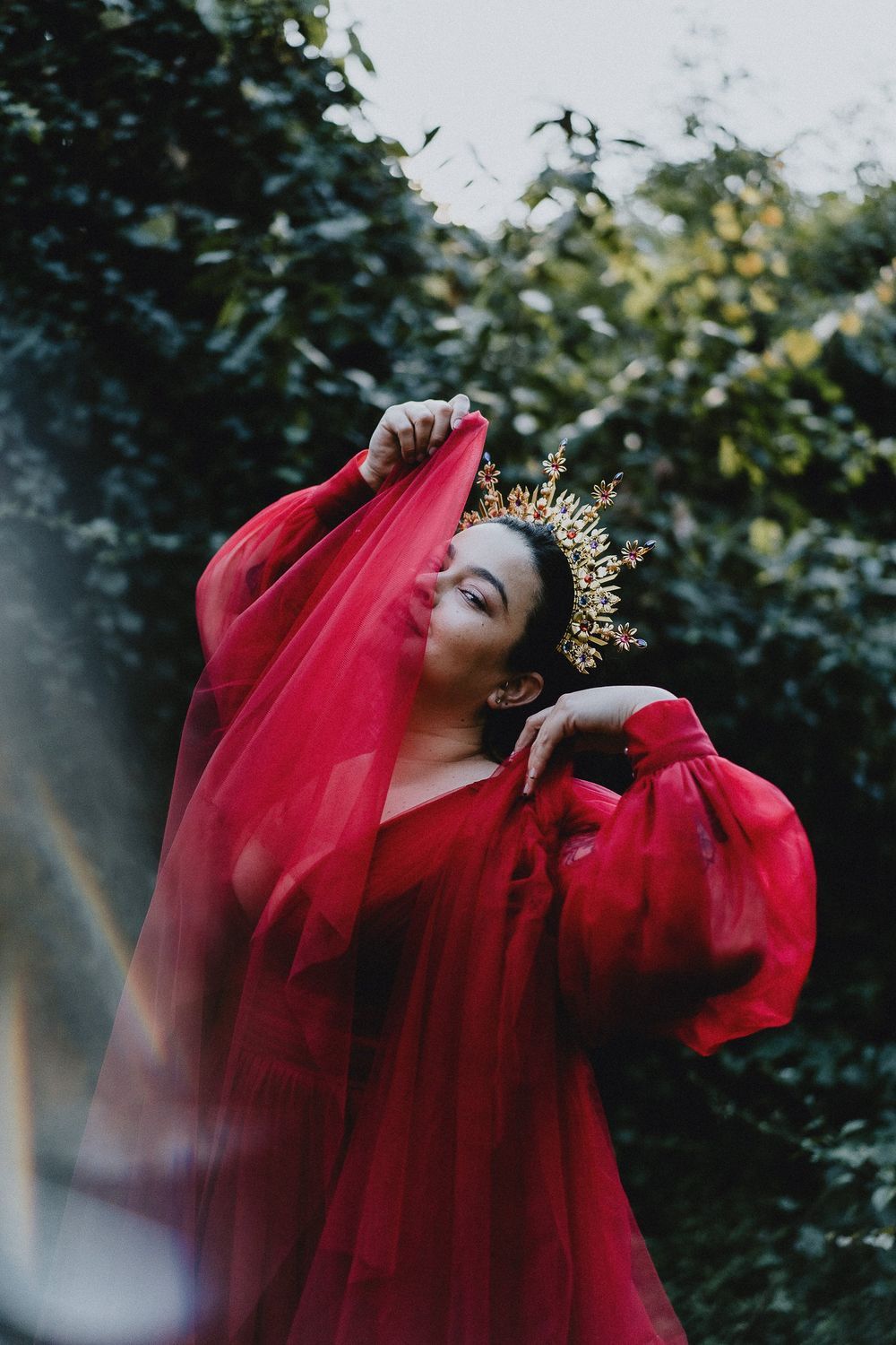 Figure in flowing red dress and golden crown poses dramatically against dark foliage background.