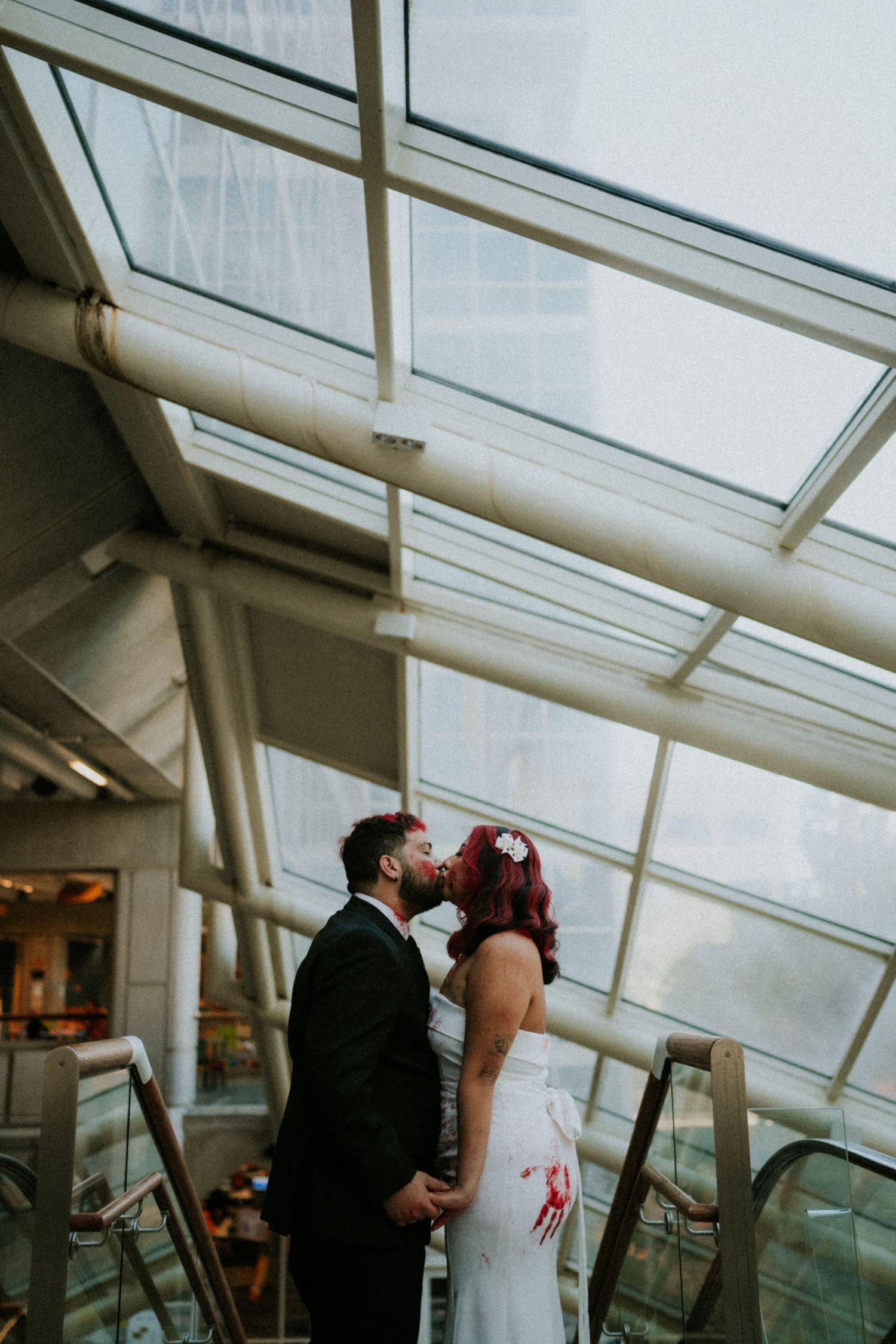Bride and groom share a romantic kiss on an escalator under a glass ceiling atrium.