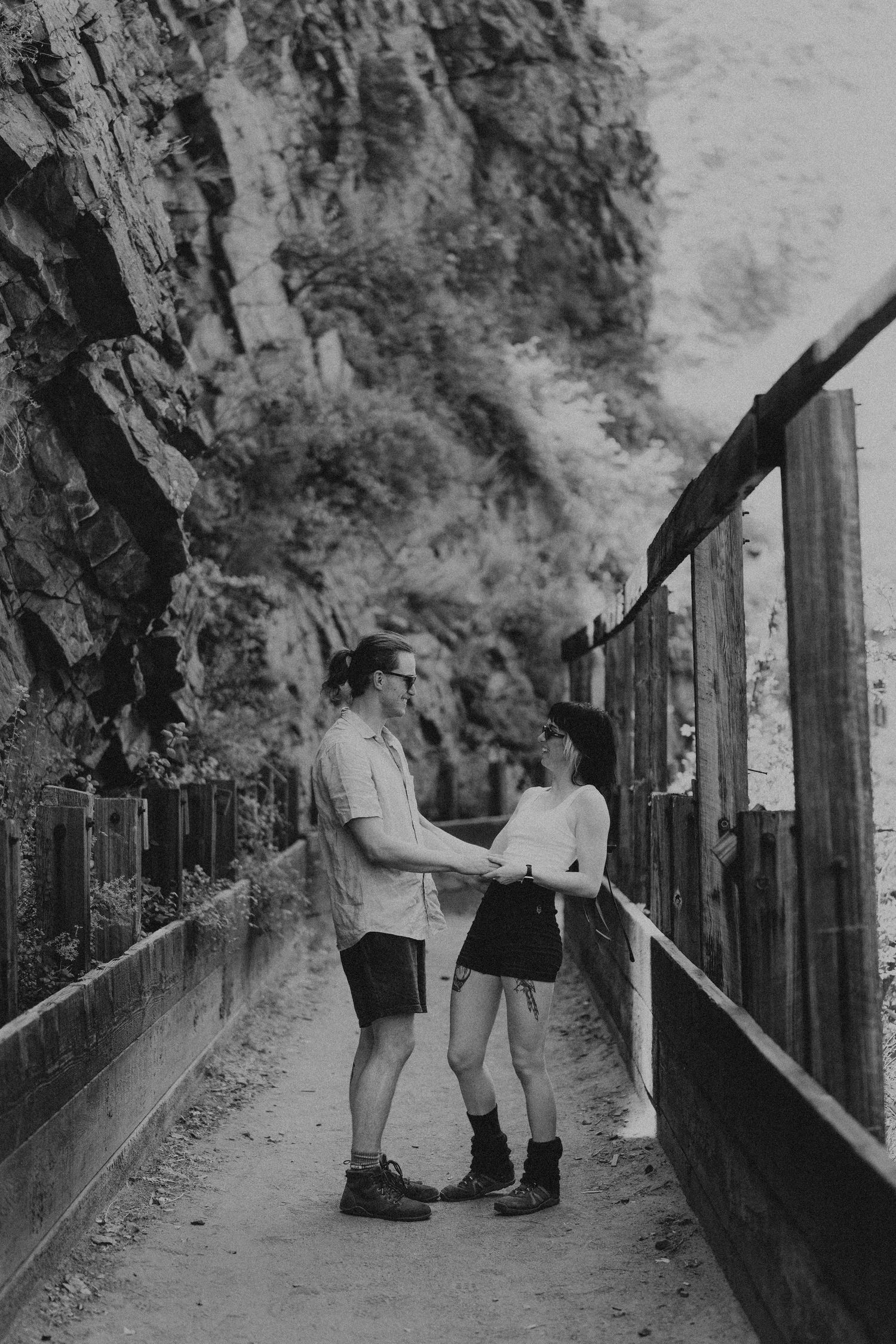 Black and white photo of a couple holding hands on a wooden walkway beside a rocky cliff.