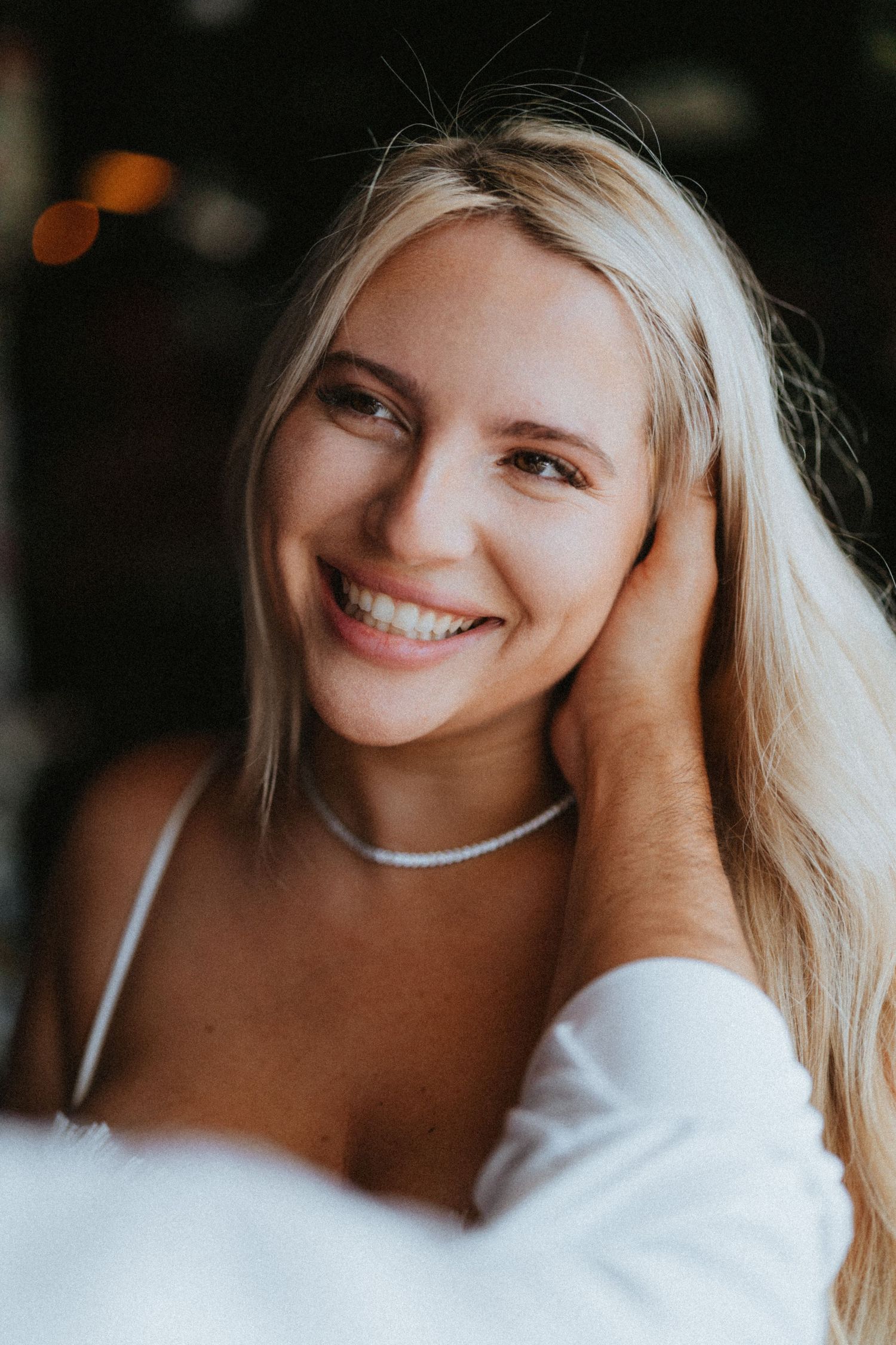 Smiling blonde woman wearing a silver necklace and white outfit, posing with hand in hair against dark background.