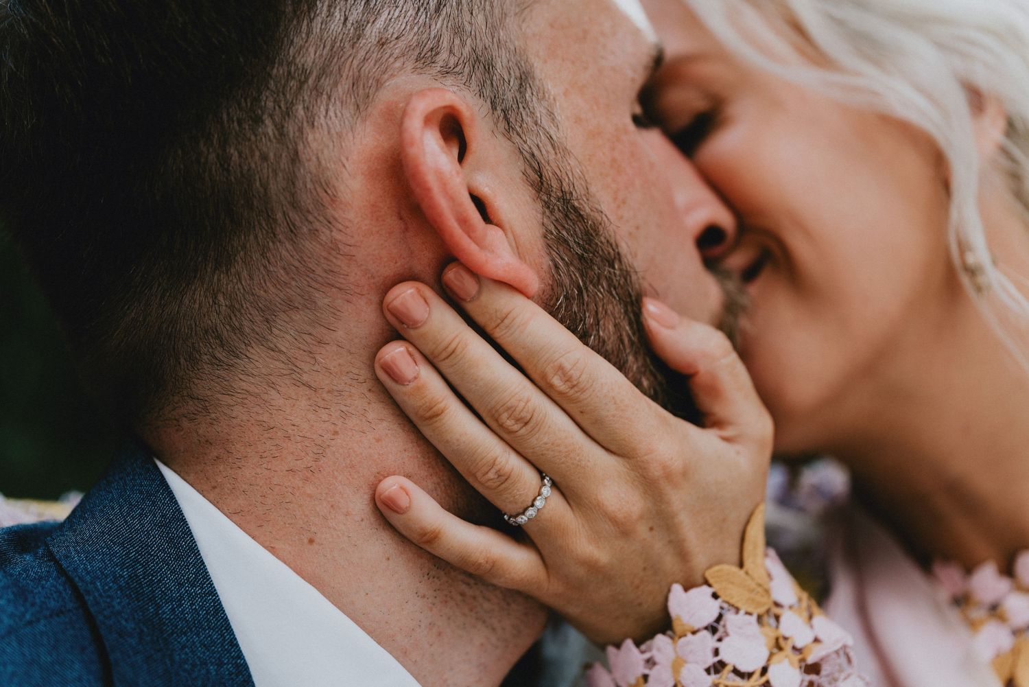 Close-up of a couple sharing a romantic kiss, woman wearing an engagement ring, man in a blue suit.