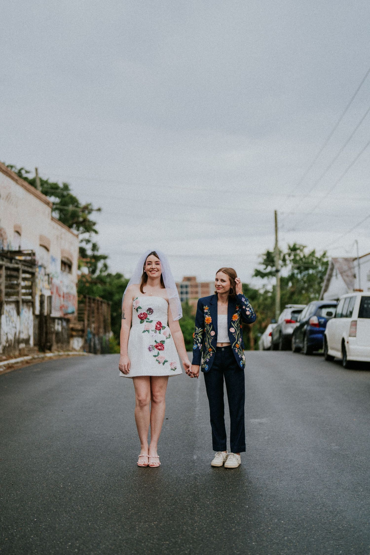 Two brides walk hand-in-hand on a street, one in a floral mini dress with veil, the other in a navy floral blazer.