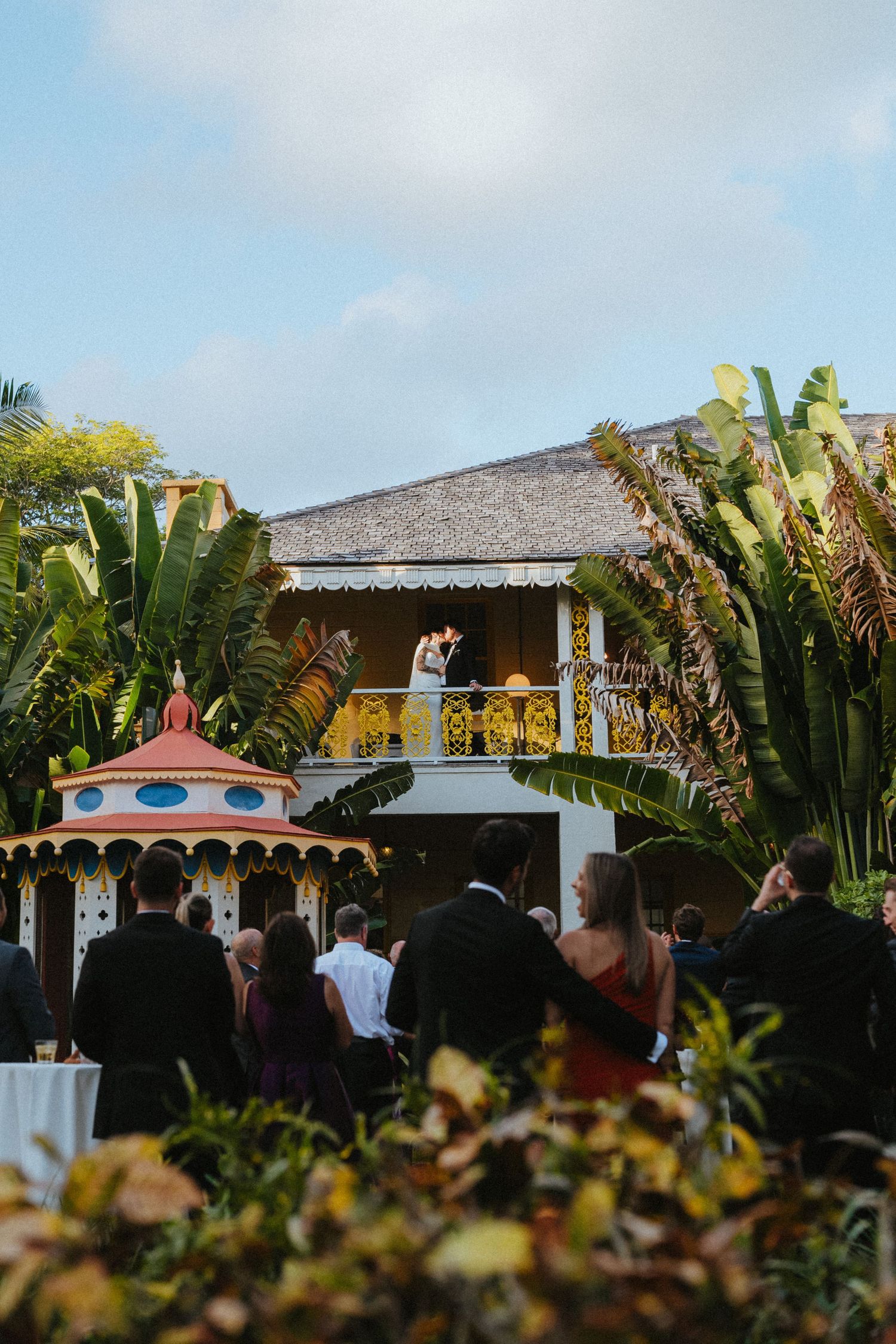 Bride and groom kiss on a yellow balcony above a colorful gazebo as guests watch below during reception.