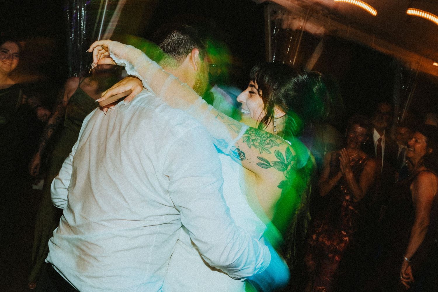 Couple dancing energetically at a wedding reception under colorful green and blue lights on a crowded dance floor.