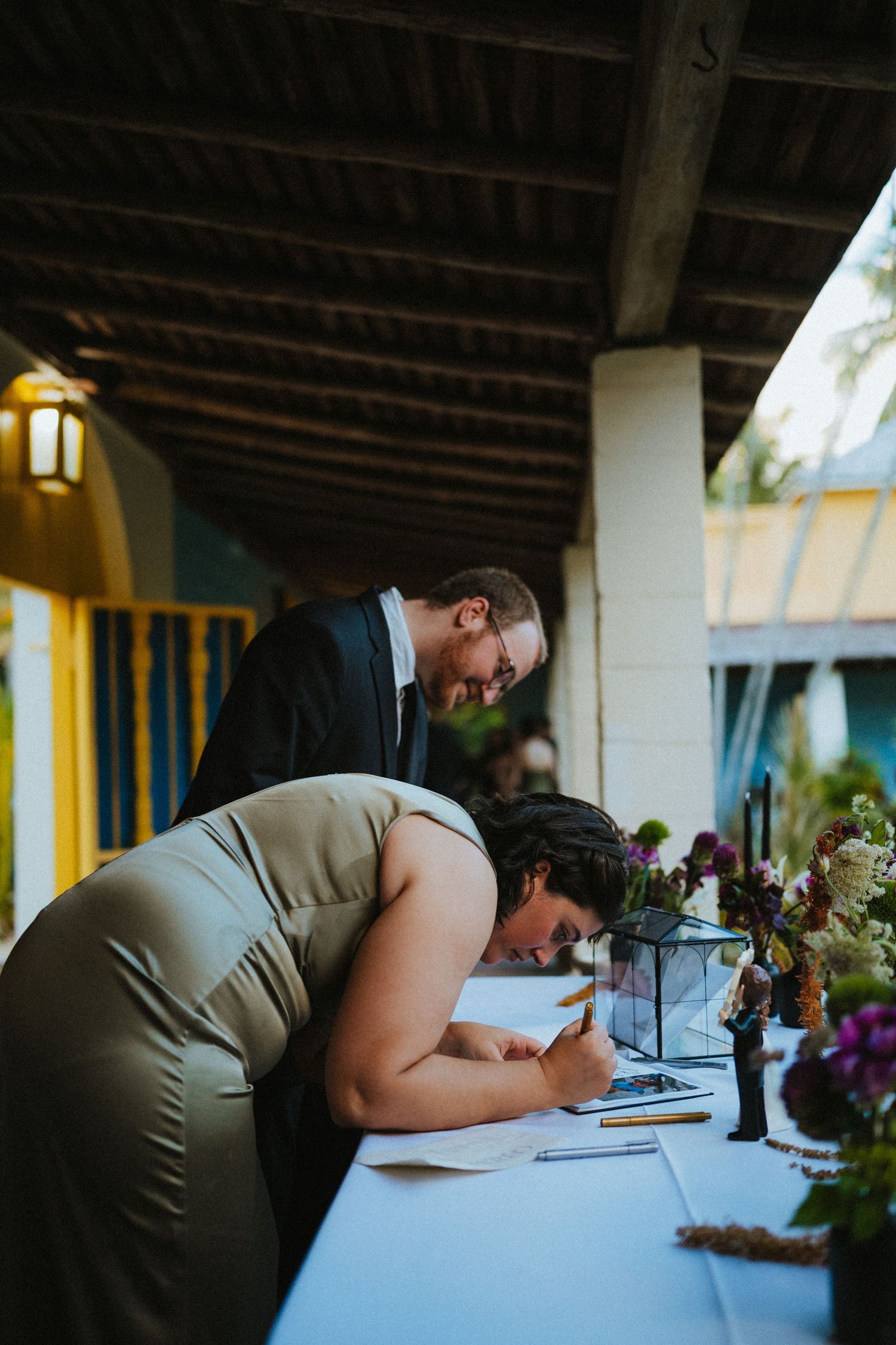 Woman in olive dress signing documents at wedding table while man in suit watches at outdoor venue.