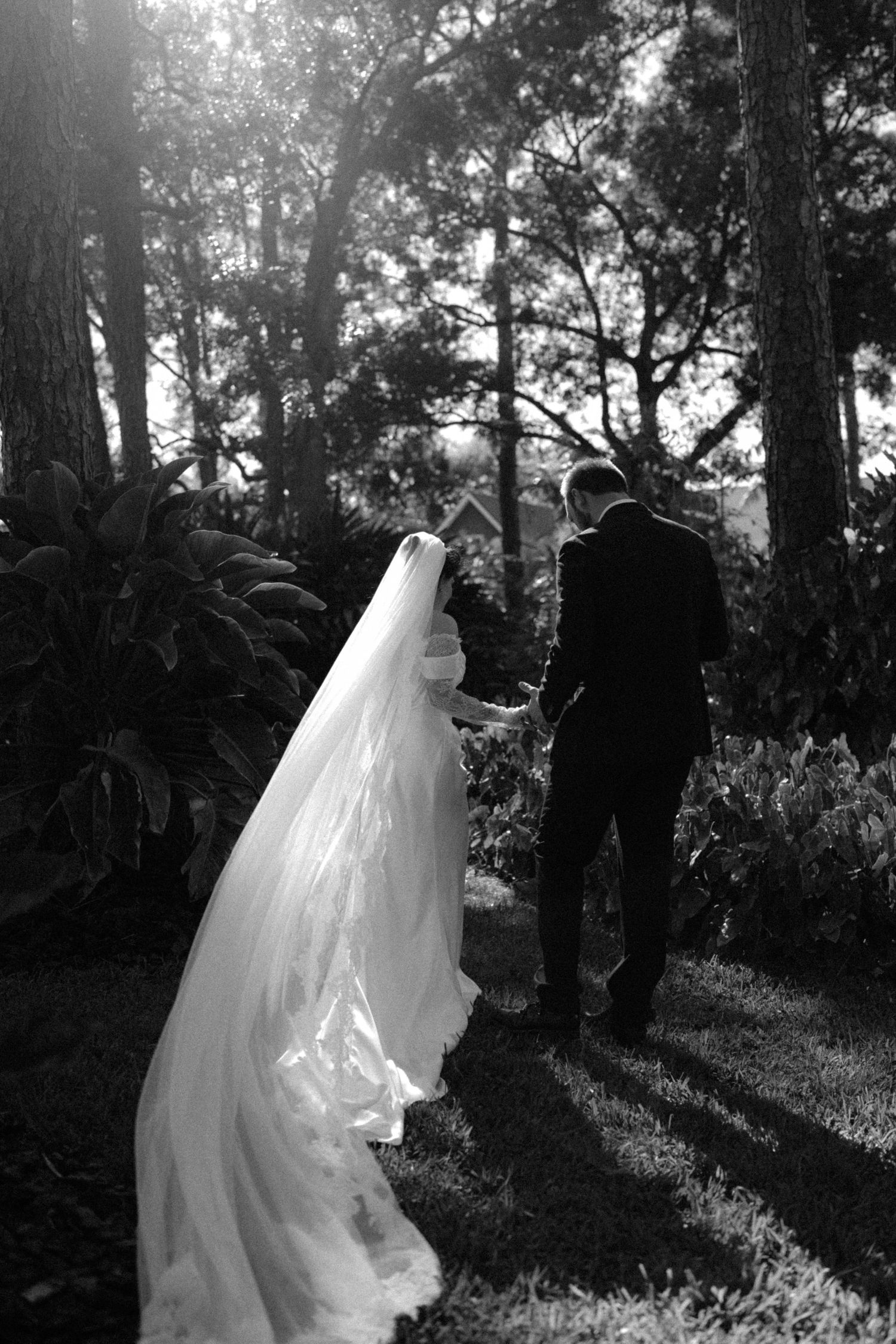 Black and white photo of bride and groom walking hand in hand through lush garden, veil trailing behind.