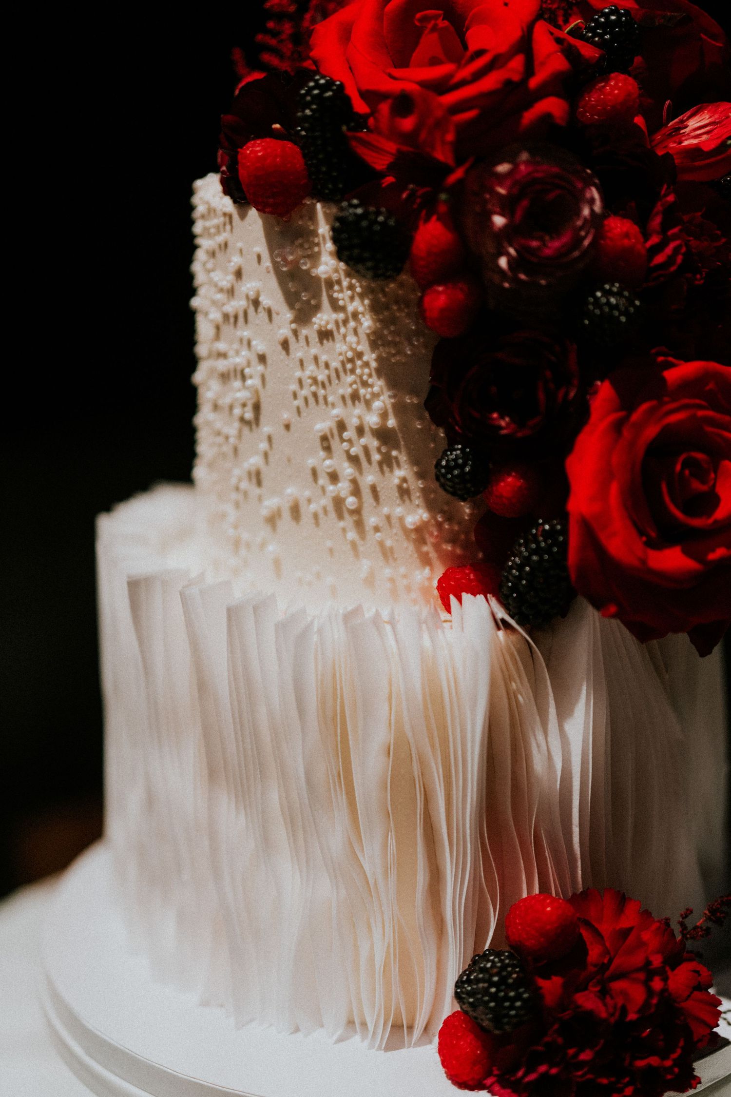 Elegant white wedding cake with red roses, blackberries, and textured ruffled fondant details.