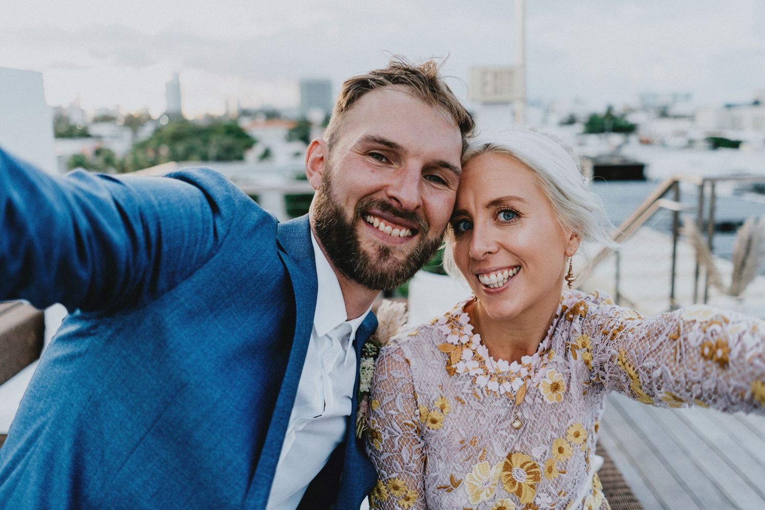 Happy couple taking a rooftop selfie, groom in blue suit and bride in floral embroidered dress on their wedding day.