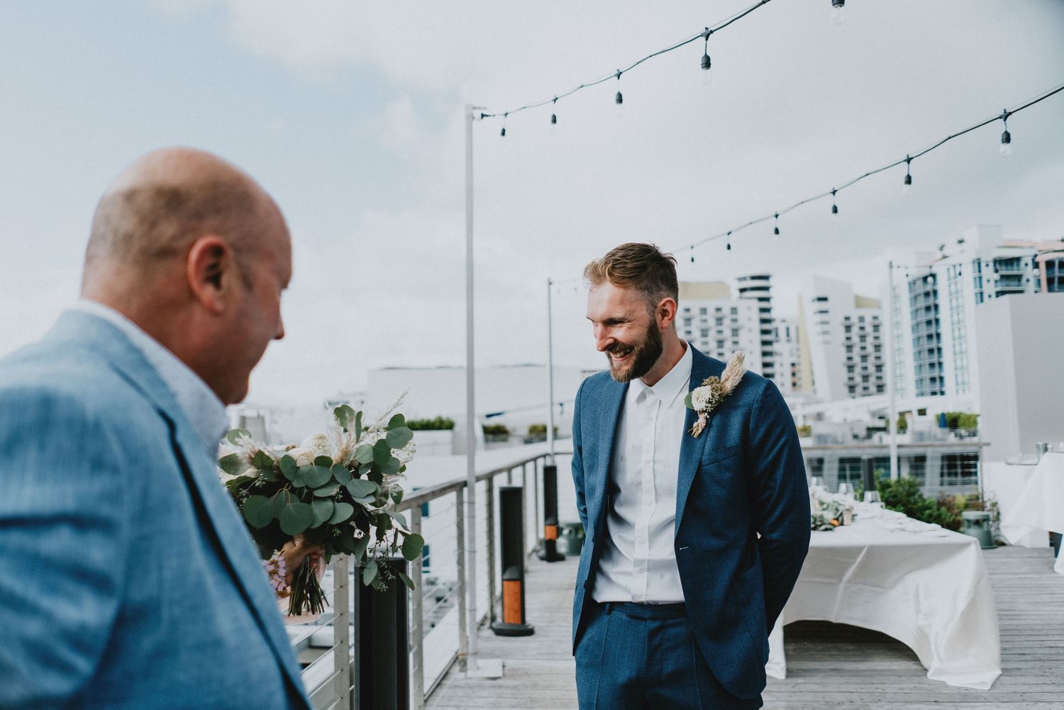 Two men in suits share a joyful moment on a rooftop wedding venue with city skyline views.