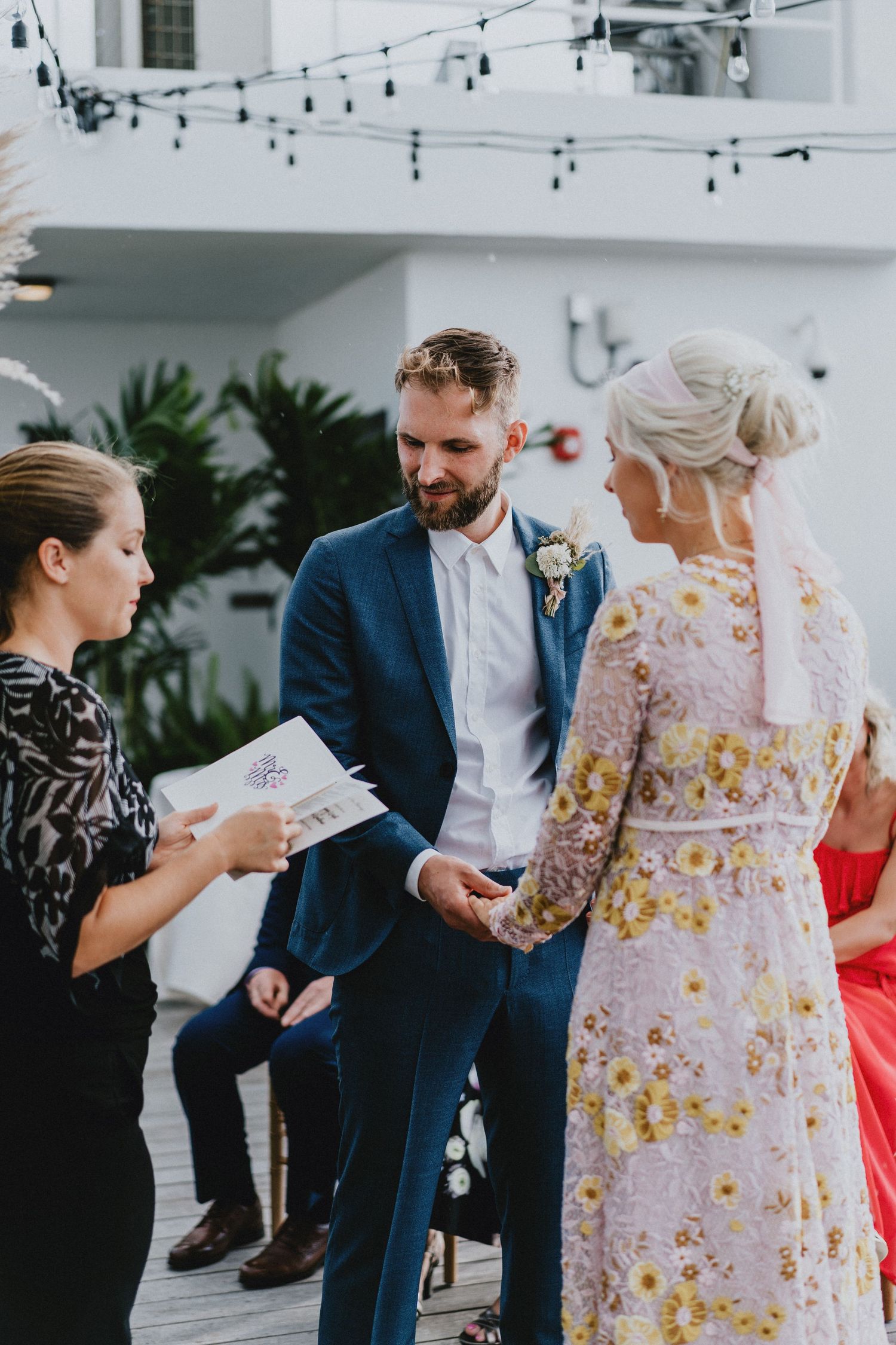 Groom in blue suit holds brides hands during outdoor wedding ceremony while officiant reads from program.