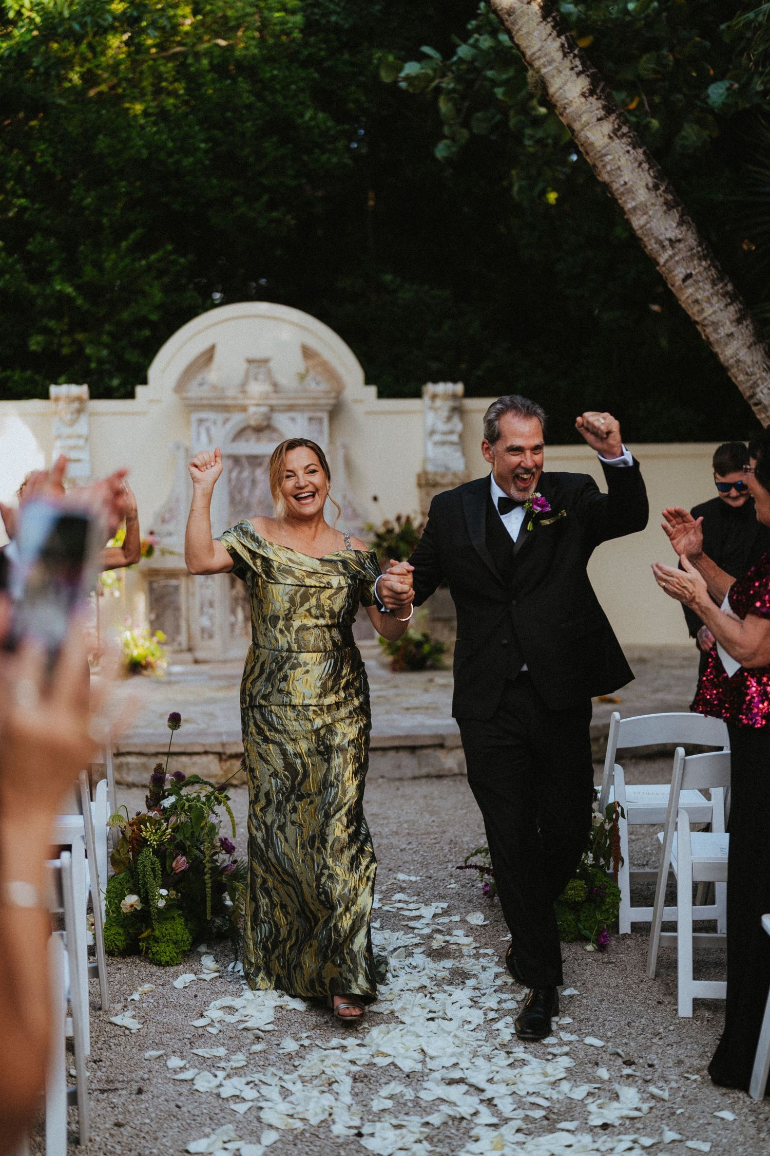 Joyful couple walks down wedding aisle as guests celebrate with confetti in an elegant outdoor garden ceremony.