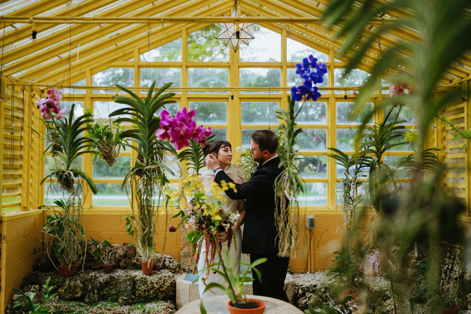 Couple embracing in a yellow greenhouse surrounded by colorful orchids and tropical plants.
