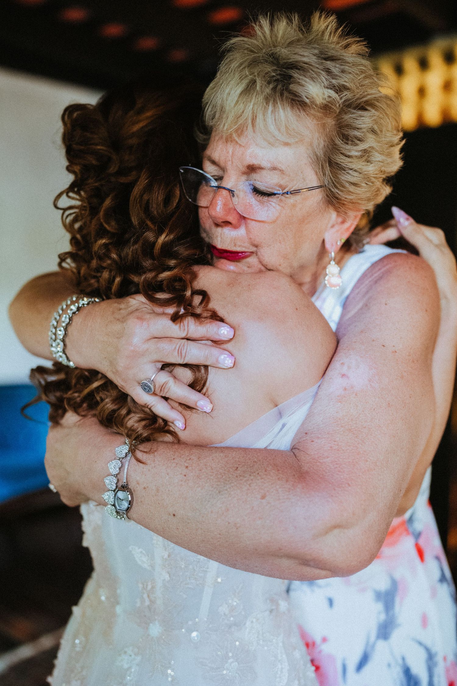 Emotional mother and bride share a heartfelt hug during wedding day preparations, both wearing elegant jewelry.