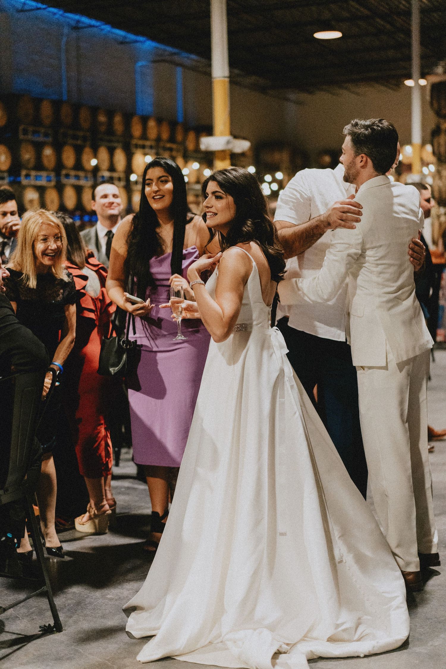 Bride in white gown smiling with groom and guests at an elegant indoor wedding reception celebration.