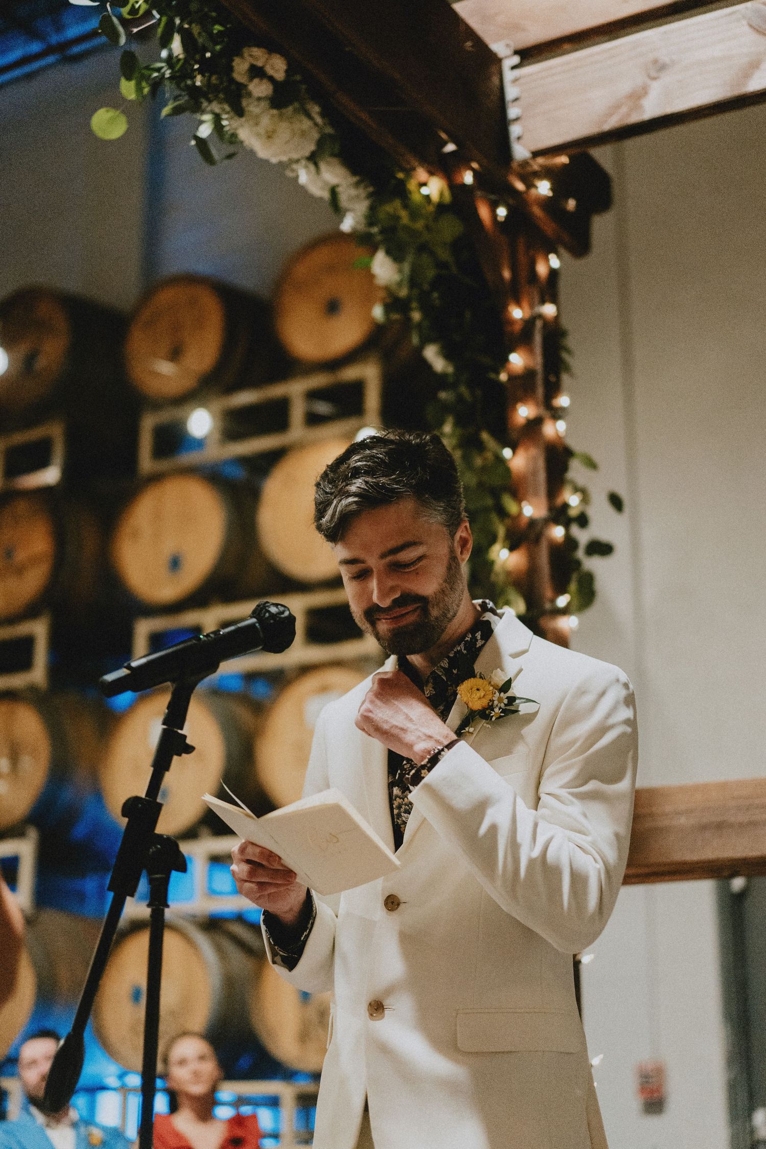 Groom in white suit reading vows at microphone with barrel wall and floral arch backdrop.