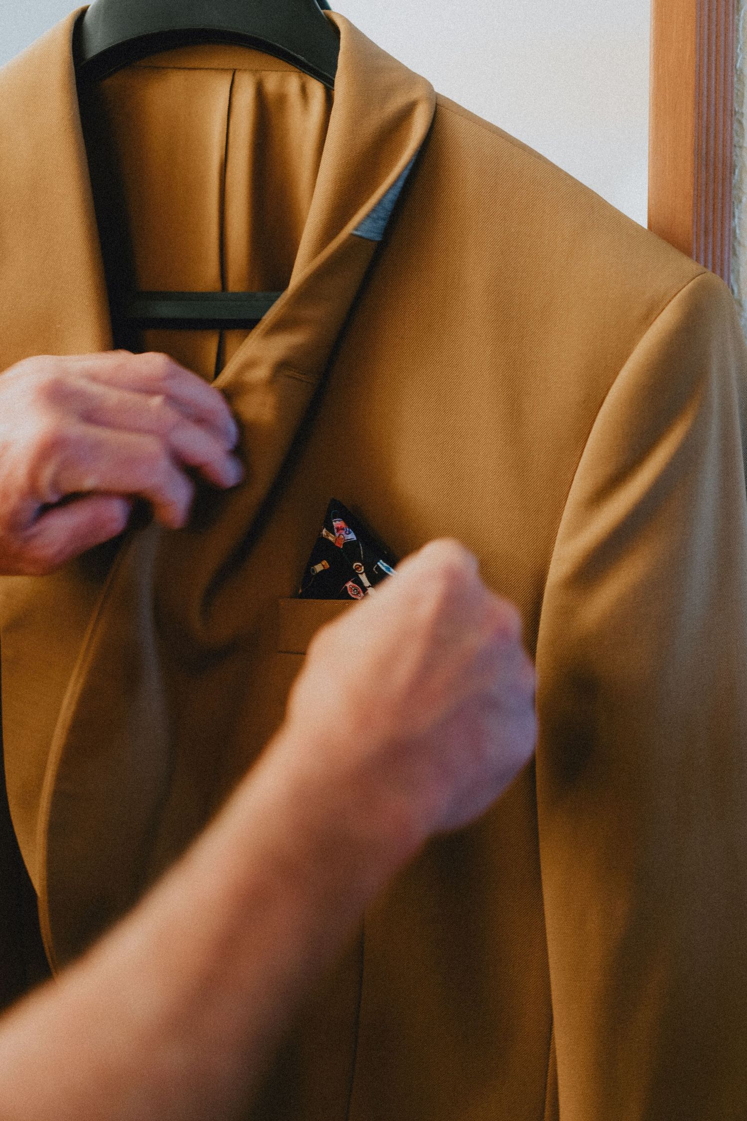 Groom adjusting pocket square on mustard yellow suit jacket hanging on hanger before wedding ceremony.