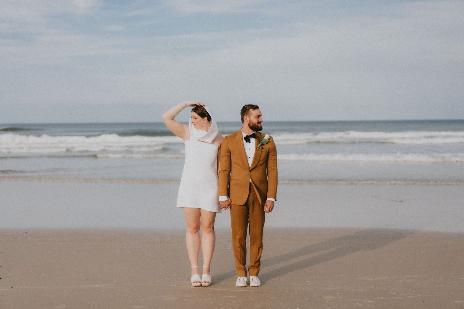 Bride and groom holding hands on sandy beach, waves crashing behind them, wearing white mini dress and brown suit.