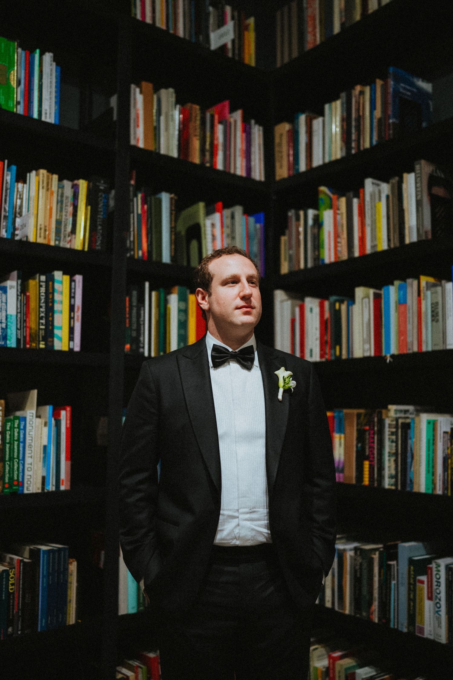 A groom in a black tuxedo with bow tie and boutonniere stands confidently between towering bookshelves.