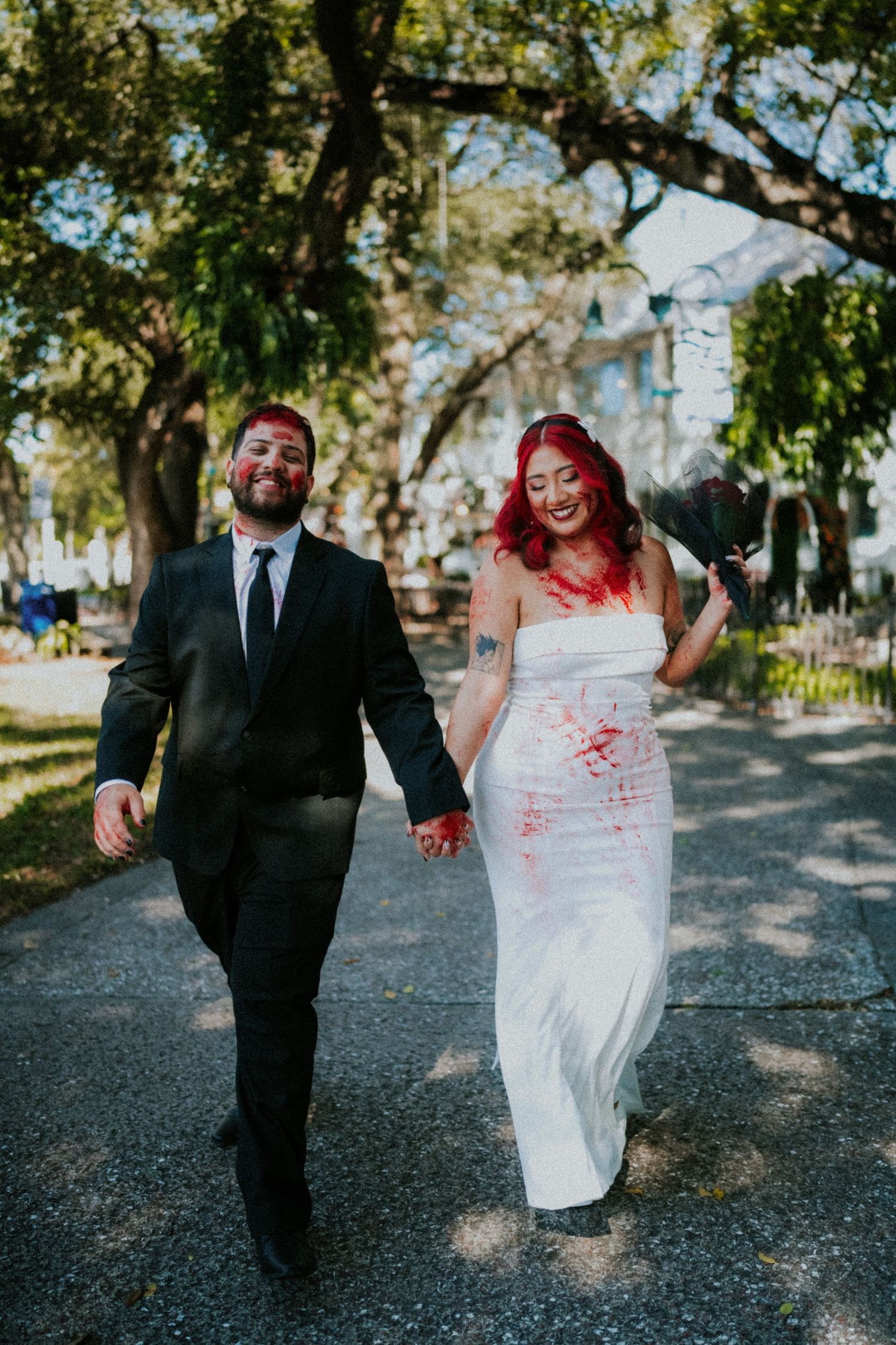 Bride in red-splattered white dress with red hair holds hands with groom in black suit walking outdoors.