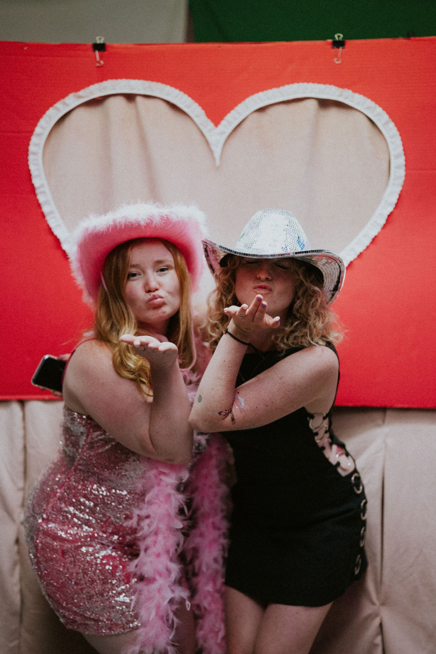 Two friends pose playfully in front of a heart backdrop wearing party dresses and cowboy hats.