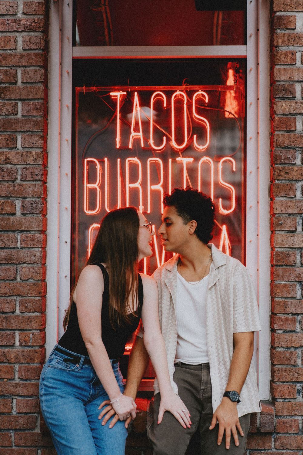 A couple stands against a brick wall with glowing red neon signs.